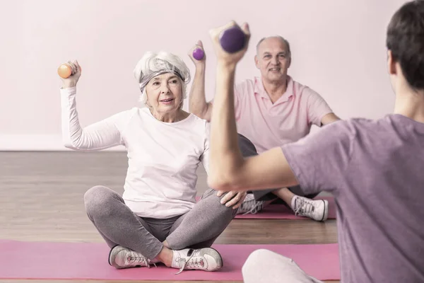 Active seniors sitting on yoga mats lifting the dumbbells during fitness classes