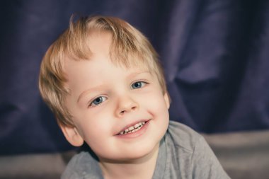 Kameraya bakıp gülümseyen sarı saçlı bir çocuğun fotoğrafı.