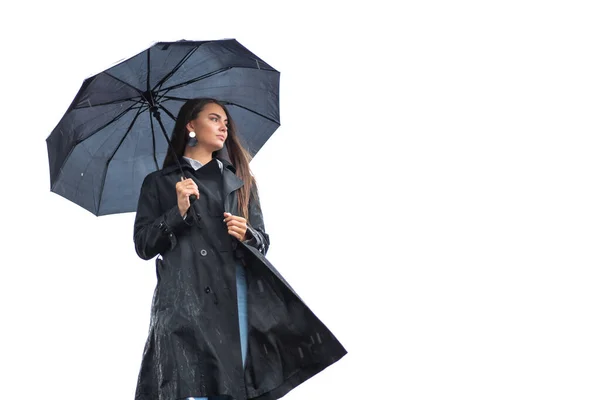 Siyah yağmurluk ve beyaz gömlekli genç bir kız yağmurlu havada beyaz bir arka planda şemsiyenin altında duruyor..