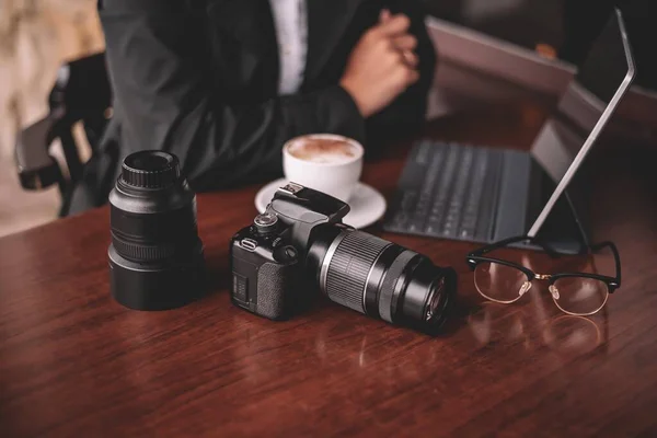 Yağmurlu havada siyah yağmurluk ve beyaz gömlekli bir kız fotoğrafçı bir fotoğraf çekiminden sonra dışarıda bir masada oturuyor kahve içiyor ve tabletteki fotoğraflara bakıyor. Masanın üzerinde bir kamera var..