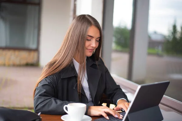Siyah yağmurluk ve beyaz gömlekli genç bir kız yağmur yağan havada bir restoranda oturuyor kahve içiyor ve tablet üzerinde daktilo yazıyor..