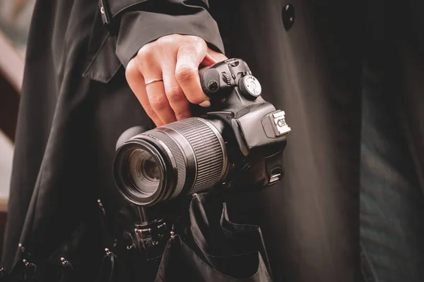 Yağmurlu havada siyah yağmurluk giyen bir fotoğrafçı kızın elinde siyah bir şemsiye ve SLR kamerası var..