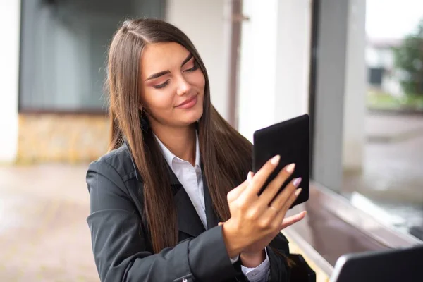 Siyah yağmurluk ve beyaz gömlekli genç bir kız yağmurlu bir havada sokakta bir restoranda oturmuş e-kitap okuyor..