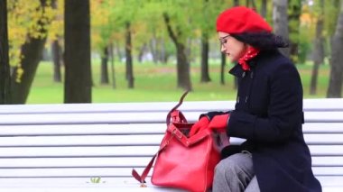 Sonbahar parkında bankta, kırmızı çanta içinde bir şey arıyor kadın.