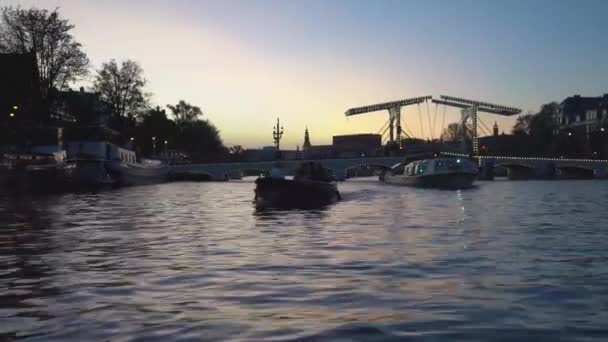 アムステルダムの夕景。運河、照明、建築、街灯で飾られた橋の下を渡るボート. — ストック動画