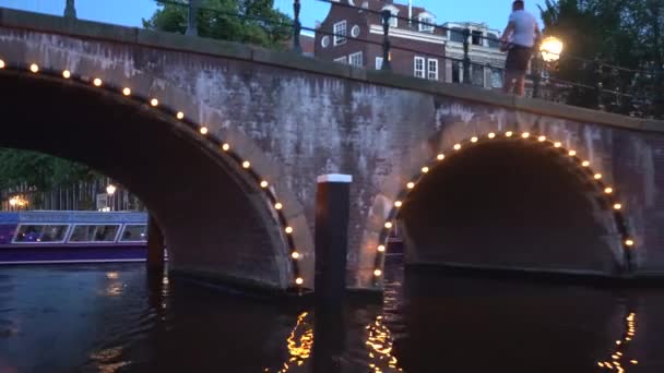 Vista serale di Amsterdam. Canali, barca che passa sotto il ponte decorata da illuminazione, architettura e lampioni . — Video Stock