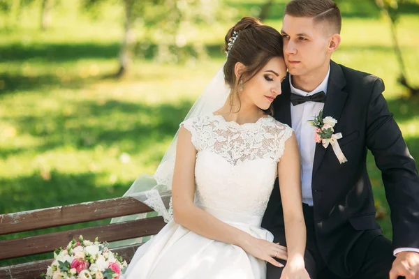 Um jovem casal de roupas de casamento está sentado em um banco em um parque. Tempo ensolarado — Fotografia de Stock