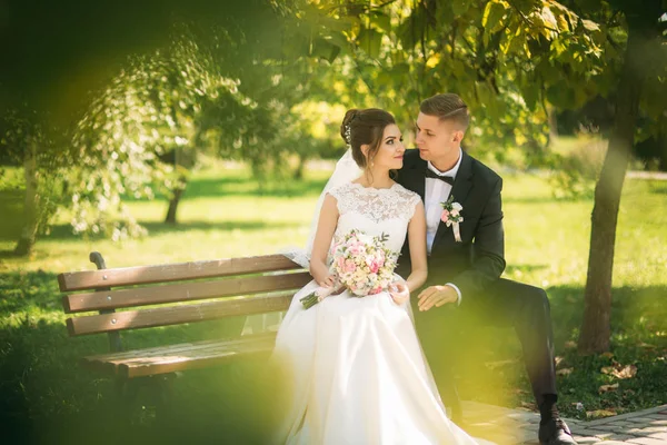 Una giovane coppia in abiti da sposa è seduta su una panchina in un parco. Tempo soleggiato — Foto Stock