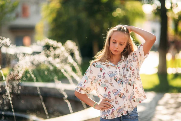 Blond tienermeisje in een blond blouse spelen met haar haren op de achtergrond van een fontein — Stockfoto
