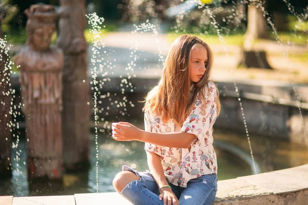 Blond teenage girl in a blond blouse playing with her hair on the background of a fountain — Stock Photo, Image