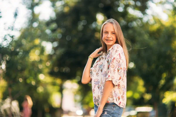 Blond flicka i blus som poserar för fotograf mot bakgrund av gröna träd — Stockfoto
