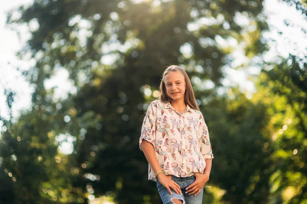 Blond dívka v halence pózuje fotografovi na pozadí Zelený strom — Stock fotografie