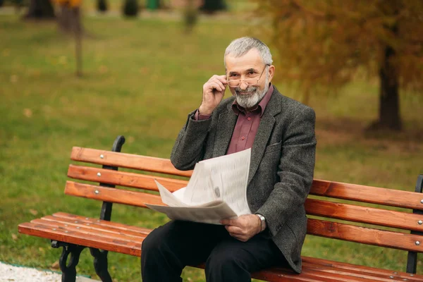 Un gentil grand-père avec une belle barbe dans une veste grise s'assoit sur un banc dans le parc et lit un journal. Loot au photographe — Photo