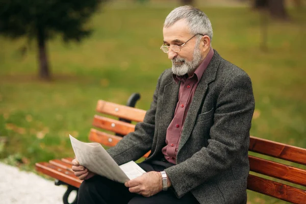 Un bel nonno con una bella barba in giacca grigia siede su una panchina nel parco e legge un giornale. — Foto Stock