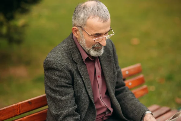 Un bel nonno con una bella barba in giacca grigia siede su una panchina nel parco e legge un giornale. — Foto Stock