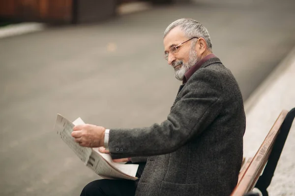 Un gentil grand-père avec une belle barbe dans une veste grise s'assoit sur un banc dans le parc et lit un journal. vue arrière — Photo