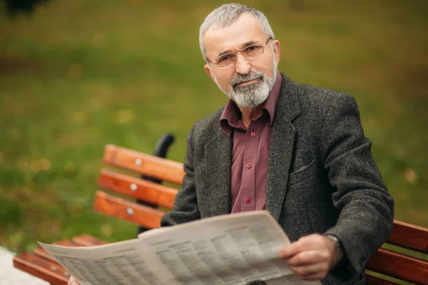 회색 자 켓에 아름 다운 수염과 좋은 할아버지 공원에서 벤치에 앉아서 신문을 읽고. 할아버지 착용 안경 — 스톡 사진