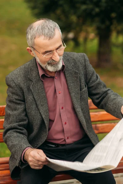 Хороший дедушка с красивой бородой в серой куртке сидит на скамейке в парке и читает газету. Дедушка носит очки — стоковое фото