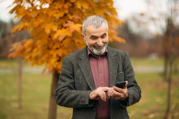 Egy jóképű idős ember visel szemüveget használ egy telefon. Őszi séta a parkban — Stock Fotó