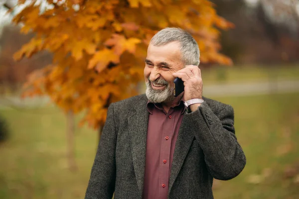 Красивый пожилой мужчина в очках пользуется телефоном. Прогулка по парку осенью — стоковое фото