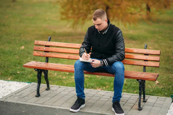 Студент в чорній куртці сидить в парку на лавці записує свої думки в блокноті. Красивий хлопчик — стокове фото