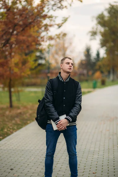 Tipo con estilo en jucket negro de pie en el parque. Tiene un cuaderno en las manos. — Foto de Stock