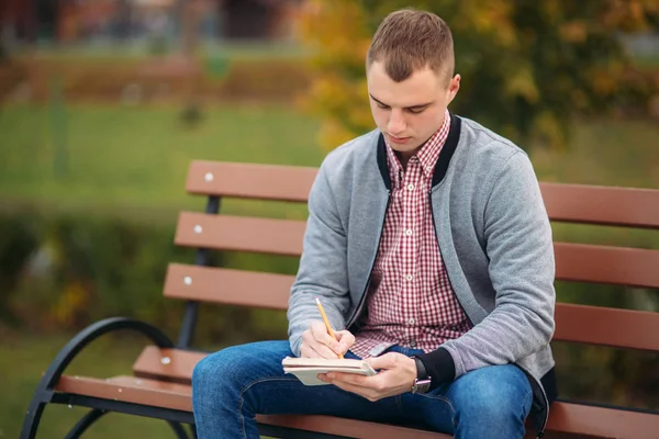 Pensil을 사용 하 여 그의 notebool에 그의 생각을 내려 벤치 쓰기에 앉아 귀여운 학생 — 스톡 사진
