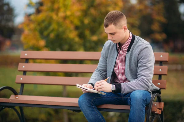 Un estudiante lindo se sienta en el banco escribe sus pensamientos en su notebool usando un pensil —  Fotos de Stock