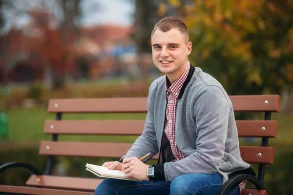 Cute student siedzi na ławce spisuje swoje myśli w jego notebool przy użyciu zarobek — Zdjęcie stockowe