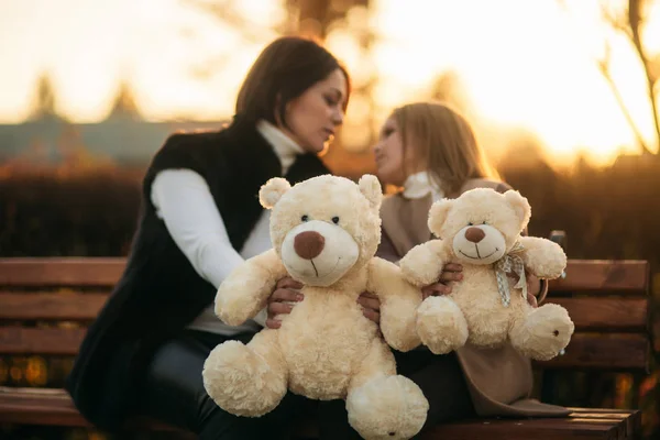 Mutter und kleine Tochter sitzen auf einer Bank im Park. hält einen Teddybär — Stockfoto