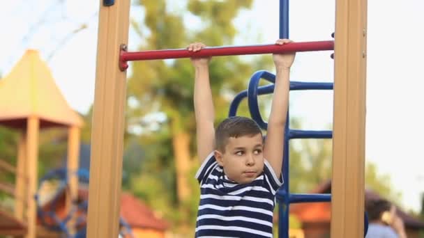 Un bambino con una maglietta a righe sta giocando nel parco giochi, oscillando su un'altalena.Primavera, tempo soleggiato — Video Stock