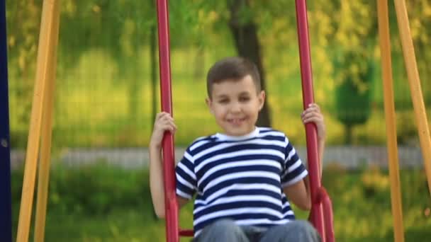 Маленький мальчик в полосатой футболке играет на детской площадке, качается на качелях. — стоковое видео