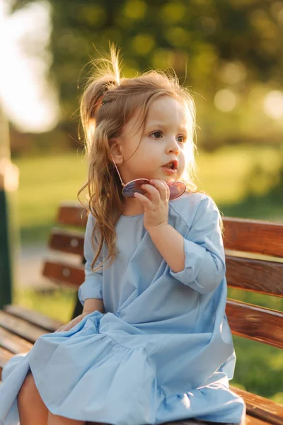 Little girl model in blue dress and sun glasses sits on a bench in the park — Stock Photo, Image
