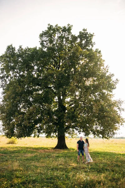 Bonito cara e uma menina atraente abraçar uns aos outros perto de uma grande árvore verde. História de amor — Fotografia de Stock