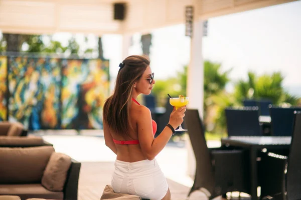 Όμορφη νεαρή γυναίκα που φοράει μαγιό πίνοντας ένα πολύχρωμο κοκτέιλ κάθεται σε μια καμπίνα από το beach bar club. Εκπληκτικό κορίτσι απολαμβάνοντας το ποτό στην θερινή ώρα — Φωτογραφία Αρχείου