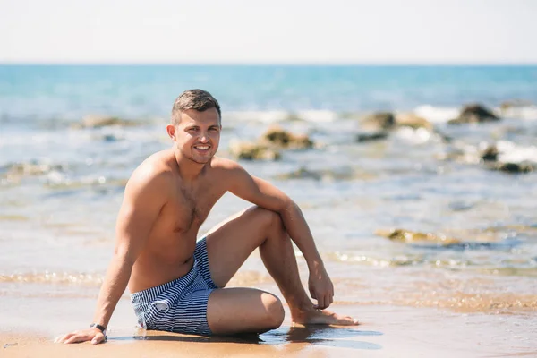 Hombre guapo en pantalones cortos sentado en la playa de arena cerca de las rocas en el fondo del mar — Foto de Stock