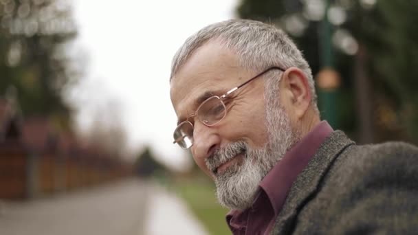 Um bom avô com uma bela barba em uma jaqueta cinza senta-se em um banco no parque e lê um jornal — Vídeo de Stock