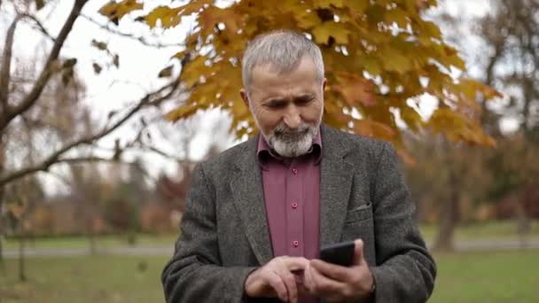 Ένας όμορφος ηλικιωμένος άνδρας φορώντας γυαλιά και χρησιμοποιώντας ένα τηλέφωνο. Βόλτα στο πάρκο το φθινόπωρο. — Αρχείο Βίντεο