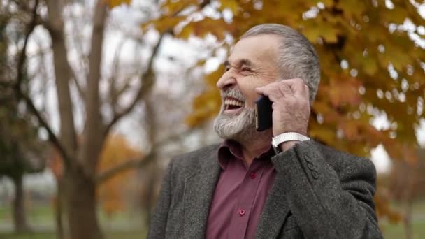 A handsome elderly man wearing glasses and using a phone. Walk in the park in autumn. — Stock Video