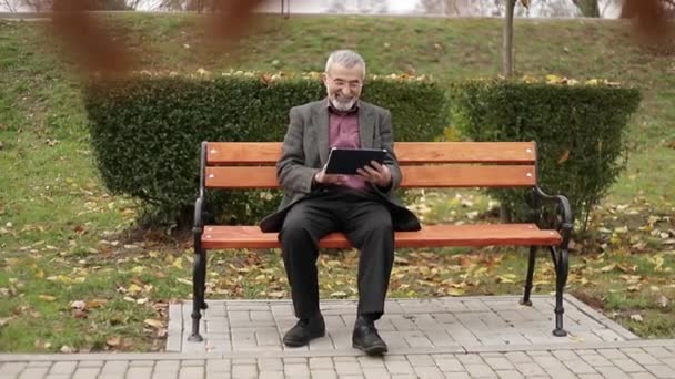 Παππούς χρησιμοποιούν ένα tablet που κάθεται στο πάρκο στον πάγκο — Αρχείο Βίντεο