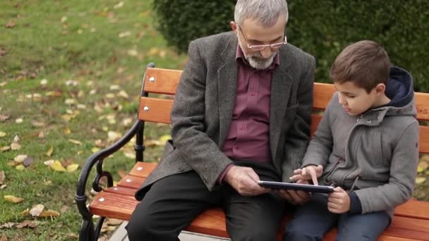 Grandpa and his grandson spend time together in the park. They are sitting on the bench and use the tablet — Stock Video