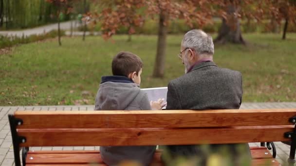 Grandpa and his grandson spend time together in the park. They are sitting on the bench and use the tablet — Stock Video