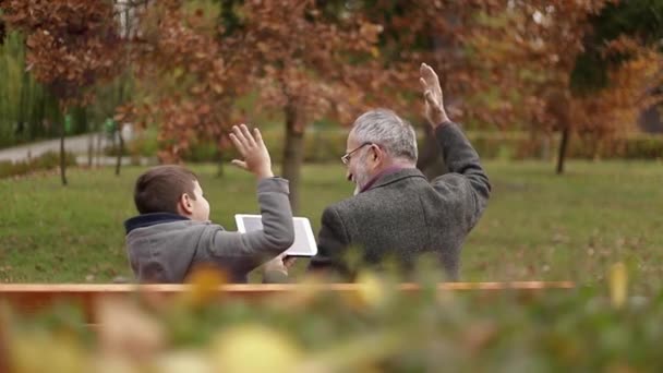 Grand-père et son petit-fils passent du temps ensemble dans le parc. Ils sont assis sur le banc et utilisent la tablette — Video