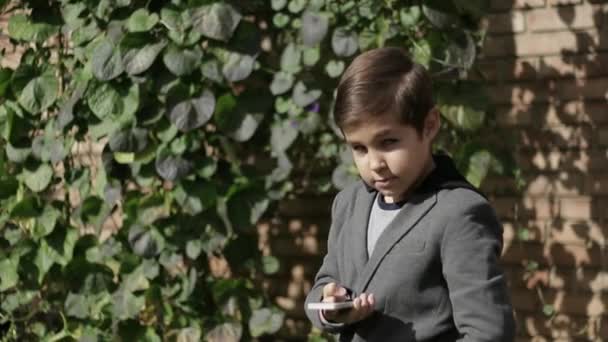 Lindo niño con chaqueta usa su teléfono. Fondo de la pared de hiedra — Vídeo de stock