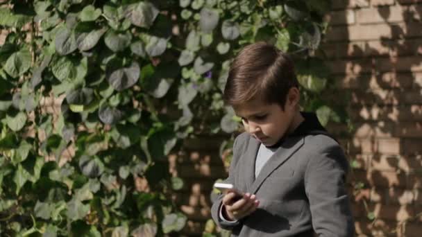 Lindo niño con chaqueta usa su teléfono. Fondo de la pared de hiedra — Vídeo de stock