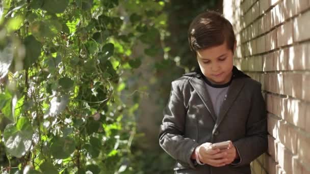 Lindo niño con chaqueta usa su teléfono. Fondo de la pared de hiedra — Vídeos de Stock