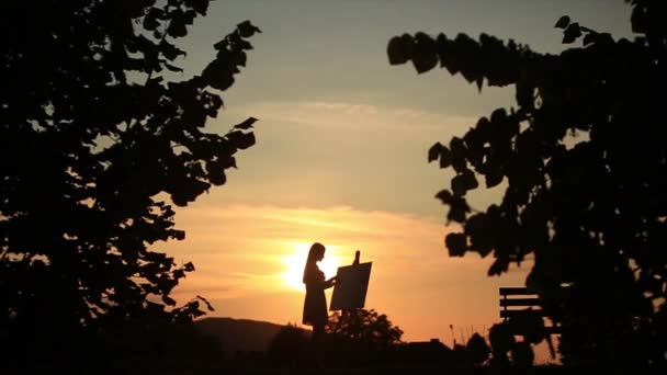 Σιλουέτα του μια ξανθιά κοπέλα ζωγραφίζει μια ζωγραφική στον καμβά με τη βοήθεια των χρωμάτων. Ένα ξύλινο καβαλέτο κρατά την εικόνα. Το καλοκαίρι είναι μια ηλιόλουστη μέρα, ηλιοβασίλεμα — Αρχείο Βίντεο