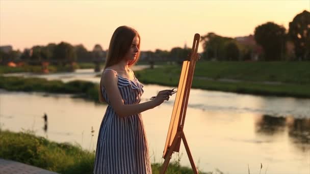 Menina bonita de pé no parque e desenha um quadro usando uma paleta com tintas e uma espátula . — Vídeo de Stock