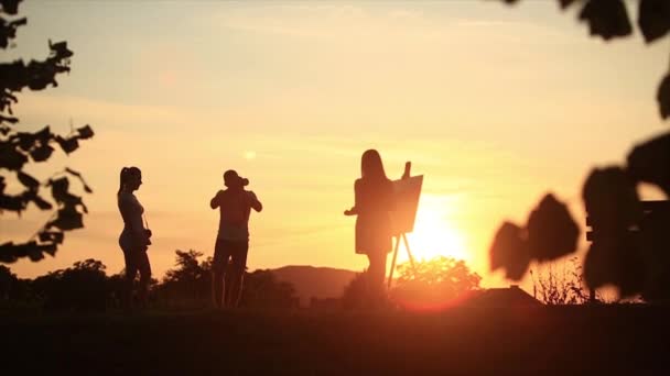 Silhouet van een blond meisje schildert een schilderij op het canvas met de hulp van verven. Een houten ezel houdt de afbeelding. De zomer is een zonnige dag, zonsondergang. Photograshers — Stockvideo