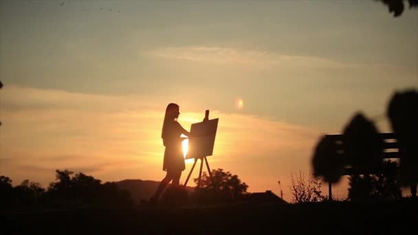 Silhouet van een blond meisje schildert een schilderij op het canvas met de hulp van verven. Een houten ezel houdt de afbeelding. De zomer is een zonnige dag, zonsondergang — Stockvideo